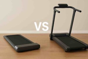 Kuvassa on kaksi erilaista juoksumattoa vierekkäin vaalean seinän ja puulattian edessä. Vasemmalla on matala ja rakenteeltaan yksinkertainen kävelymatto ilman kaidetta. Oikealla on korkeampi, perinteinen juoksumatto, jossa on tukikaiteet ja ohjauspaneeli. Kuvan keskellä on suuri “VS”-teksti, joka korostaa vertailuasetelmaa.