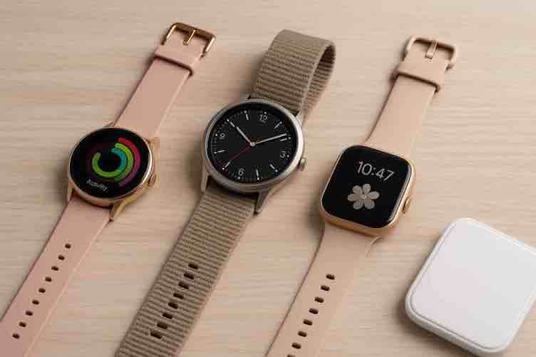 Kolme erilaista älykelloa on aseteltu vierekkäin vaalealle puupinnalle. Vasemmalla on pyöreänäyttöinen kultareunainen kello vaaleanpunaisella hihnalla ja värikkäällä aktiivisuusnäkymällä. Keskellä on perinteisen kellon näköinen malli tummalla analogisella kellotaululla ja hiekanvärisellä kudotulla hihnalla. Oikealla on neliömäinen kulta-reunainen älykello beigeillä hihnoilla ja näytöllä kukkakuviolla sekä kellonajalla 10:47.