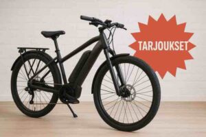 Musta sähköpyörä seisoo vaalealla lattialla valkoista tiiliseinää vasten. Pyörän oikealla puolella on suuri oranssi, räjähdysmuotoinen grafiikka, jossa lukee ”TARJOUKSET”. Kuvassa erottuvat pyörän runko, akku ja etupyörän levyjarru.
