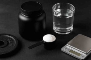 Mustalla pöydällä on musta kreatiinipurkki, sen edessä mittalusikallinen valkoista jauhetta, lasillinen vettä sekä pieni digitaalinen vaaka. Vasemmassa alakulmassa näkyy myös painolevy. Esineet on aseteltu siististi tummalle taustalle.
