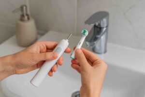 Kaksi kättä pitää valkoista sähköhammasharjaa ja irrotettavaa harjaspäätä kylpyhuoneen lavuaarin edessä. Vasemman käden sormet tukevat laitetta, kun taas oikea käsi kohdistaa pyöreäharjaksisen pään kiinnitysvarteen. Taustalla näkyy metallinen hana ja saippuapumppu.